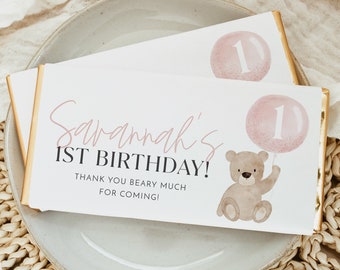 May include: A white chocolate bar with a pink and gold design. The bar has a pink balloon with the number 1 on it and a brown teddy bear holding the balloon. The text on the bar reads "Savannah's 1st Birthday! Thank you beary much for coming!"