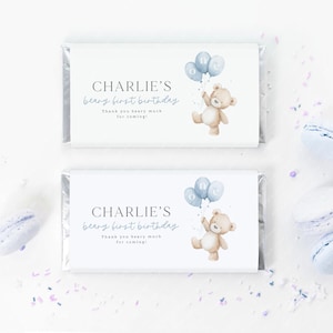 May include: Two white candy bar wrappers with text and illustrations. The text reads "CHARLIE'S beary first birthday" and "Thank you beary much for coming!". A teddy bear holds blue balloons. The wrappers are decorated with blue confetti.