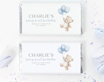 May include: Two white candy bar wrappers with text and illustrations. The text reads "CHARLIE'S beary first birthday" and "Thank you beary much for coming!". A teddy bear holds blue balloons. The wrappers are decorated with blue confetti.