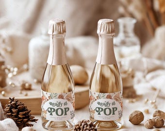 Puede incluir: Dos pequeñas botellas de champán con etiquetas que dicen "Listo para estallar" y presentan un tema de bosque con setas, hojas y animales. Las botellas están sobre una superficie marrón claro con piñas y nueces.