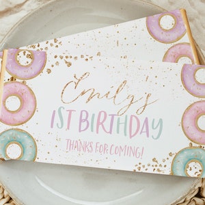 Może przedstawiać: Białe pudełko upominkowe z złotym brokatem i różowymi i niebieskimi ilustracjami pączków. Tekst na pudełku brzmi "Emily's 1st Birthday Thanks for Coming!"