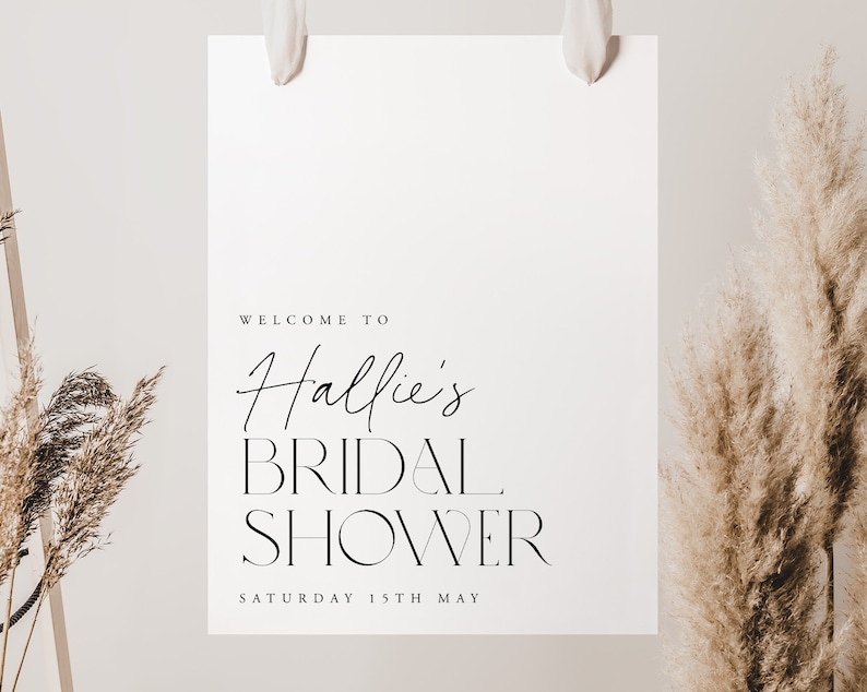 May include: A white sign with the text "WELCOME TO Hallie's BRIDAL SHOWER SATURDAY 15TH MAY" in black script and block letters. The sign is hanging from white ribbons. Dried ornamental grasses are on either side of the sign.