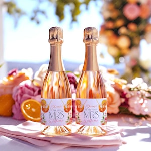 Könnte beinhalten: Zwei Champagnerflaschen mit goldfarbenen Folienverschlüssen und Etiketten mit der Aufschrift "Cheers! To the Future MRS Williams" mit einer Cocktail-Illustration. Die Flaschen sind mit einer rosa Flüssigkeit gefüllt und stehen auf einem rosa Tuch, umgeben von Blumen und einer Orangenscheibe.