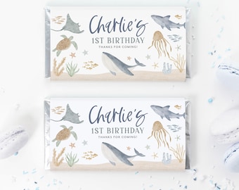 May include: Two white chocolate bar wrappers with a blue and brown ocean theme. The wrappers feature a whale, stingray, turtle, jellyfish, and coral. The text on the wrappers reads "Charlie's 1st Birthday Thanks for coming!"