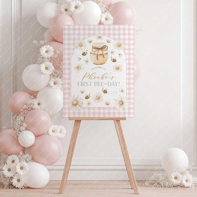 May include: A welcome sign for a first birthday celebration, featuring a pink gingham background and floral accents. The sign reads "Welcome to Phoebe's First Bee-Day!" and is displayed on a wooden easel, surrounded by balloons and flowers.