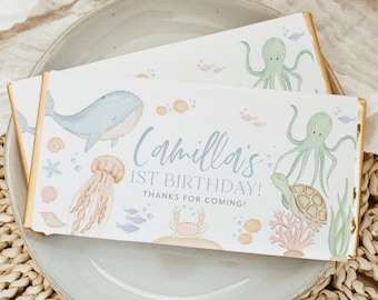 May include: A white chocolate bar with a blue whale, orange jellyfish, green octopus, and a turtle. The text on the bar reads "Camilla's 1st Birthday! Thanks for coming!"