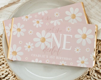 May include: Two pink chocolate bar wrappers with a daisy pattern and gold edges. The wrappers have the text "isn't she ONE derful" and "Charlie's 1st Birthday". The bars are on a light gray plate and a woven placemat.