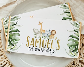 May include: A white chocolate bar wrapper with a jungle theme. It features a giraffe, elephant, rhino, and lion with green leaves and a gold foil accent. The text reads "Samuel's 1st birthday!"