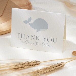 Op de afbeelding: Een gevouwen witte kaart met een lichtblauwe walvisillustratie. De kaart toont "THANK YOU" in donkergrijs, met "Love Samantha + Jackson" eronder. De kaart ligt op een wit oppervlak, vergezeld van tarwearen.