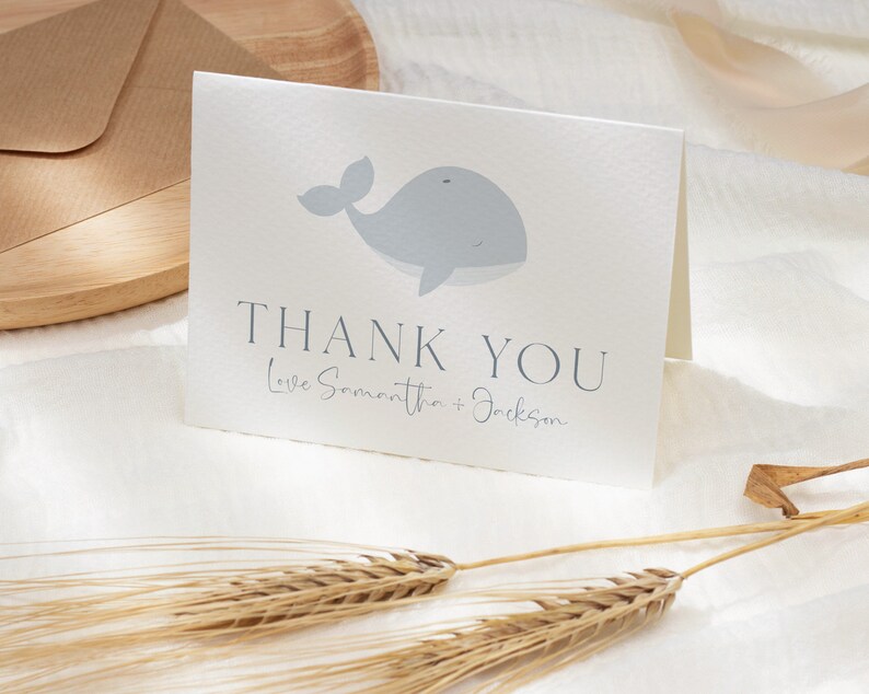 Op de afbeelding: Een gevouwen witte kaart met een lichtblauwe walvisillustratie. De kaart toont "THANK YOU" in donkergrijs, met "Love Samantha + Jackson" eronder. De kaart ligt op een wit oppervlak, vergezeld van tarwearen.