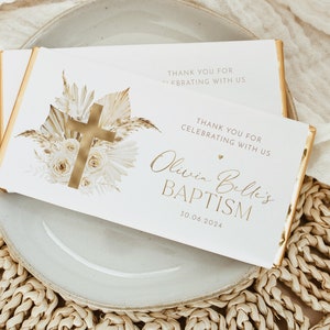 May include: Two white chocolate bar wrappers with gold accents and text. One wrapper features a gold cross and floral design. The other wrapper says "Olivia Belle's BAPTISM 30.06.2024" and "THANK YOU FOR CELEBRATING WITH US."