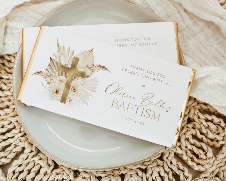 May include: Two white chocolate bar wrappers with gold accents and text. One wrapper features a gold cross and floral design. The other wrapper says "Olivia Belle's BAPTISM 30.06.2024" and "THANK YOU FOR CELEBRATING WITH US."