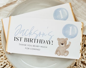 May include: A white chocolate bar with gold foil wrapping. The bar has a blue and white watercolor design with a teddy bear holding a blue balloon with the number 1 on it. The text on the bar reads "Jackson's 1st Birthday! Thank you beary much for coming!"