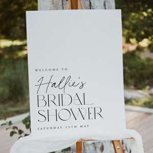 May include: A white sign on an easel with the text "Welcome to Hallie's Bridal Shower Saturday 15th May" in black script and block letters. The sign is draped with white fabric and set outdoors.