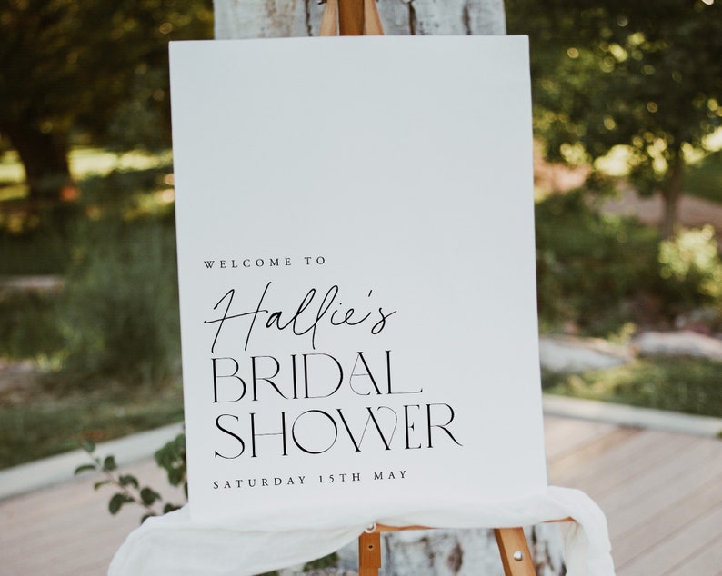 May include: A white sign on an easel with the text "Welcome to Hallie's Bridal Shower Saturday 15th May" in black script and block letters. The sign is draped with white fabric and set outdoors.