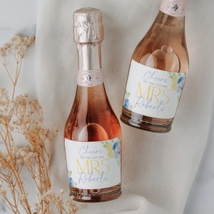May include: Two mini bottles of pink sparkling wine with personalized labels. The labels feature a watercolor floral design with blue and yellow flowers. The text on the labels reads "Cheers to the future Mrs. Roberts".