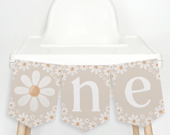 Op de afbeelding: Een beige slinger met het woord "one" in witte letters, versierd met witte madeliefjes en lichtbruine centra. De slinger is ontworpen voor een eerste verjaardag en is bevestigd aan een witte kinderstoel.