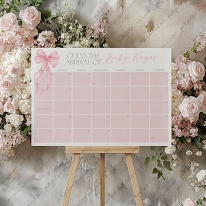 Pode incluir: Um calend&aacute;rio rosa com um la&ccedil;o no topo e o texto "Adivinhe a chegada do Baby Hayes". O calend&aacute;rio est&aacute; em um cavalete de madeira e tem os meses de abril e maio.