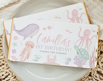 May include: A pink and purple watercolor design on a white chocolate bar wrapper. The wrapper features a whale, octopus, turtle, jellyfish, and crab. The text on the wrapper reads "Camilla's 1st Birthday! Thanks for coming!"