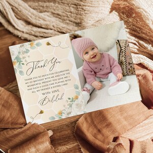 May include: A thank you card with a floral and honeycomb design. The card features a photo of a baby girl wearing a pink hat and sweater. The text on the card reads "Thank You" and "Thank you so much for celebrating my 1st birthday with me and for the wonderful present you gave me! Had such a good time playing with all my friends & eating cake! With love, Delilah."
