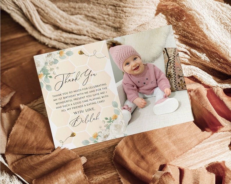 May include: A thank you card with a floral and honeycomb design. The card features a photo of a baby girl wearing a pink hat and sweater. The text on the card reads "Thank You" and "Thank you so much for celebrating my 1st birthday with me and for the wonderful present you gave me! Had such a good time playing with all my friends & eating cake! With love, Delilah."