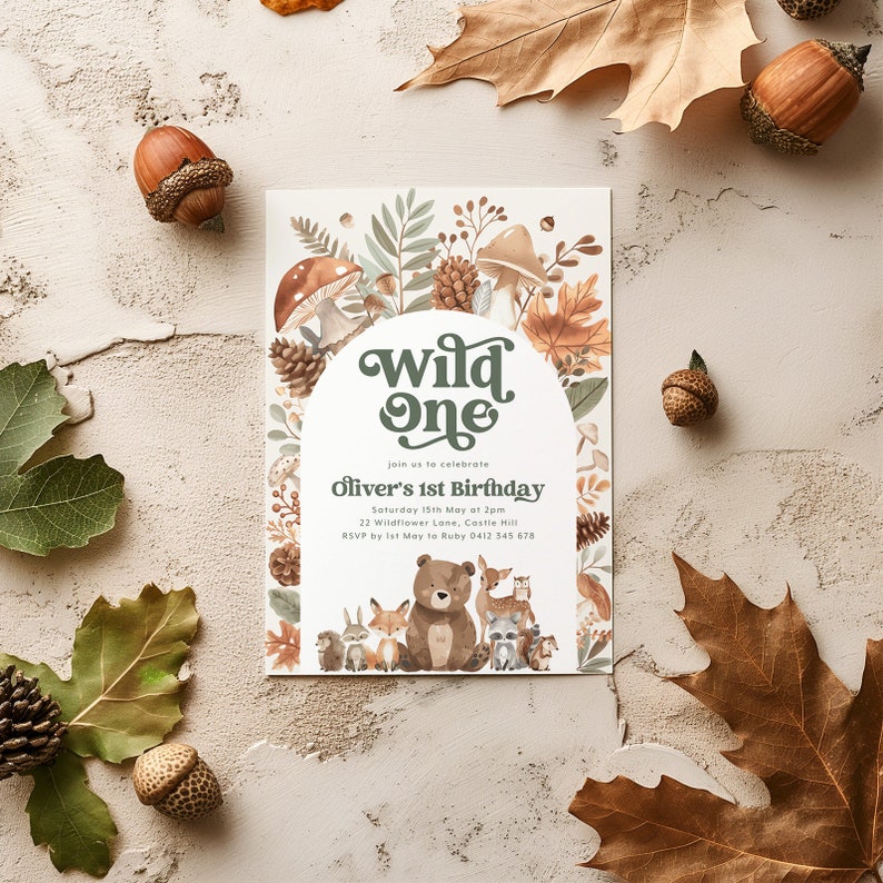 May include: A white invitation card with a woodland theme. The card features a green and brown arch with the words "wild one" in a retro font. The invitation is for Oliver's 1st birthday party. The card is decorated with illustrations of woodland animals, mushrooms, and leaves.