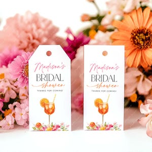 Könnte beinhalten: Zwei weiße Geschenkanhänger mit dem Text "Madison's BRIDAL shower THANKS FOR COMING!" in rosa Schrift. Jeder Anhänger zeigt eine Aquarellillustration eines Cocktails mit einer Orangenscheibe und floralen Akzenten. Oben ist ein Loch gestanzt.