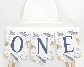 May include: A blue and white high chair banner with the word "ONE" in large blue letters. The banner features a watercolor design of sea creatures, including whales, sharks, and starfish.
