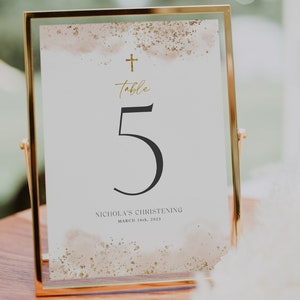 May include: A gold-framed table number card with a watercolor background and gold glitter accents. The number 5 is printed in black and the text "Table" is written in gold script. The text "Nicholas's Christening March 16th, 2023" is printed in black at the bottom.