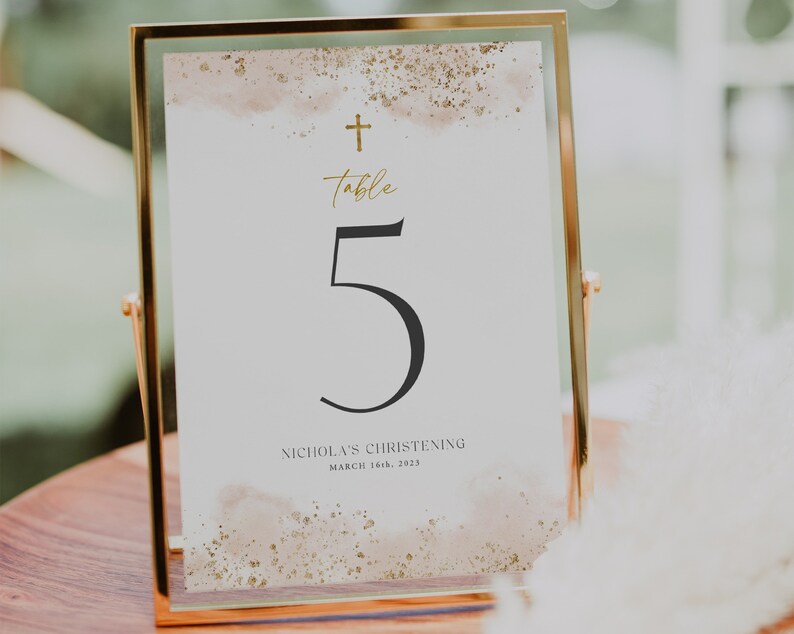 May include: A gold-framed table number card with a watercolor background and gold glitter accents. The number 5 is printed in black and the text "Table" is written in gold script. The text "Nicholas's Christening March 16th, 2023" is printed in black at the bottom.