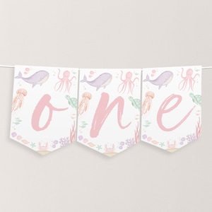 Può includere: Uno striscione rosa e bianco con la parola "one" scritta in corsivo rosa. Lo striscione è decorato con un motivo a tema marino con balene, meduse, polpi e tartarughe marine.