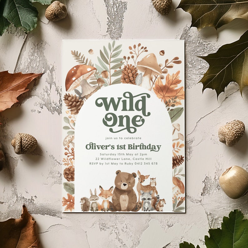 May include: A woodland-themed first birthday party invitation with a green and brown "Wild One" banner, surrounded by illustrations of mushrooms, leaves, and pine cones. The text reads "Join us to celebrate Oliver's 1st Birthday, Saturday 15th May at 2pm, 22 Wildflower Lane, Castle Hill, RSVP by 1st May to Ruby 0412 345 678."
