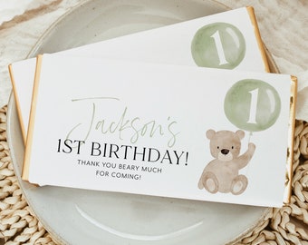 May include: A white chocolate bar wrapper with a green and brown illustration of a teddy bear holding a green balloon with the number "1" on it. The text "Jackson's 1st Birthday! Thank you beary much for coming!" is printed on the wrapper.