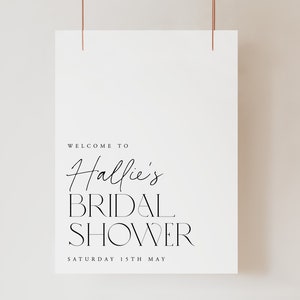 May include: A white poster with the text "WELCOME TO Hallie's BRIDAL SHOWER SATURDAY 15TH MAY" in black script and sans-serif fonts. The poster is suspended by two thin, copper-colored wires.