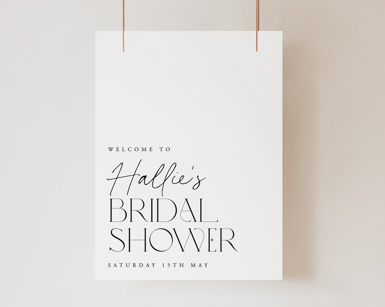 May include: A white poster with the text "WELCOME TO Hallie's BRIDAL SHOWER SATURDAY 15TH MAY" in black script and sans-serif fonts. The poster is suspended by two thin, copper-colored wires.