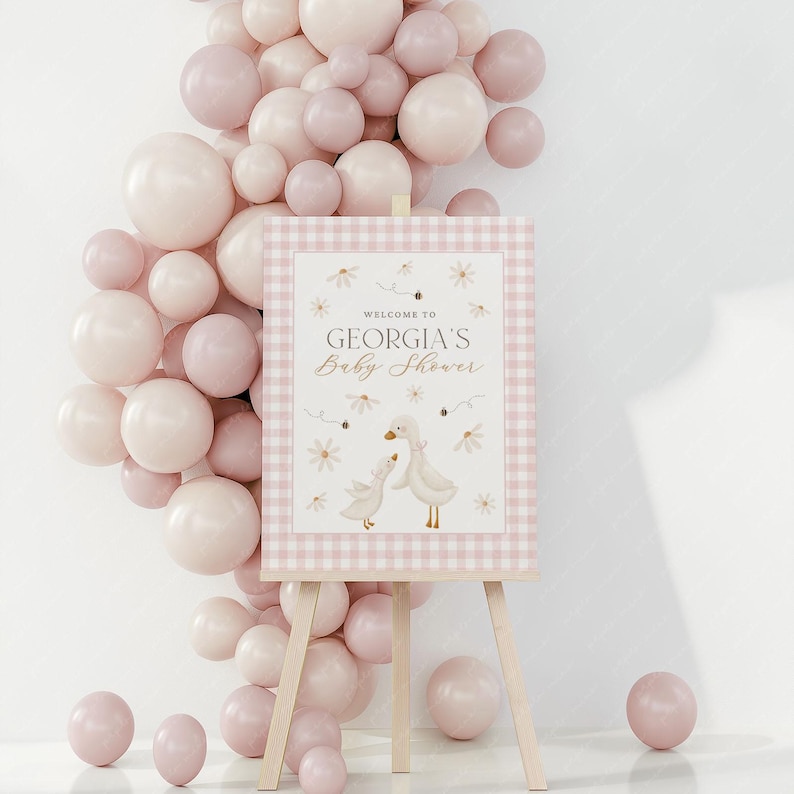 May include: A pink and white gingham baby shower sign with a welcome message and a cute illustration of two geese. The sign is on an easel in front of a wall decorated with pink balloons.