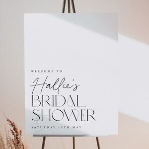 May include: A white sign on a wooden easel with the words "WELCOME TO Hallie's BRIDAL SHOWER" in black script and serif fonts. The date "SATURDAY 15TH MAY" is printed below. The sign is in front of a neutral background.