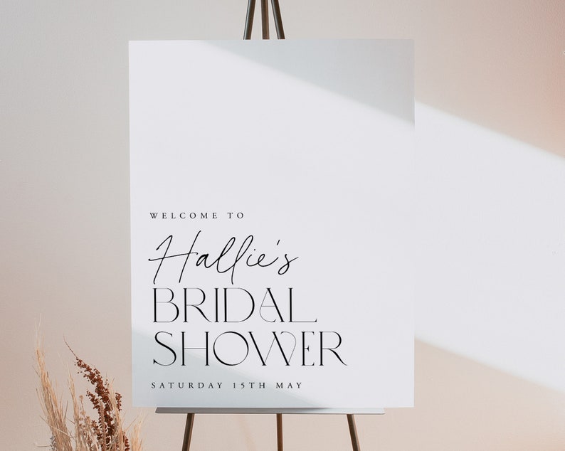 May include: A white sign on a wooden easel with the words "WELCOME TO Hallie's BRIDAL SHOWER" in black script and serif fonts. The date "SATURDAY 15TH MAY" is printed below. The sign is in front of a neutral background.