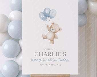 May include: A sign for a first birthday celebration. The sign features a watercolour illustration of a teddy bear holding blue balloons that spell "one." The text reads "welcome to CHARLIE'S beary first birthday Saturday 12th May."