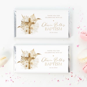 May include: Two white chocolate bar wrappers with gold foil accents. Each wrapper features a gold cross, floral design, and text that reads "THANK YOU FOR CELEBRATING WITH US" and "Olivia Belli's BAPTISM 30.06.2024". Pink and white macarons are scattered around.