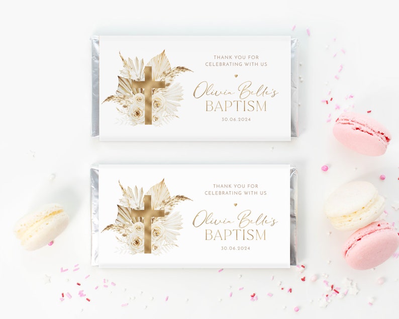 May include: Two white chocolate bar wrappers with gold foil accents. Each wrapper features a gold cross, floral design, and text that reads "THANK YOU FOR CELEBRATING WITH US" and "Olivia Belli's BAPTISM 30.06.2024". Pink and white macarons are scattered around.