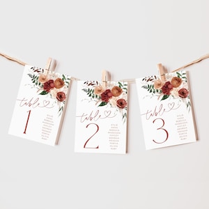 Puede incluir: Tres tarjetas de mesa blancas con diseños florales y las palabras "mesa 1", "mesa 2" y "mesa 3" en texto marrón. Cada tarjeta enumera un grupo de nombres en texto marrón.
