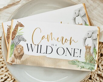 May include: White chocolate bar with gold foil accents and a jungle animal theme. The bar has a white background with the text "Cameron is a Wild One!" in gold lettering. The bar is decorated with illustrations of a giraffe, zebra, lion, elephant, crocodile, and leopard.