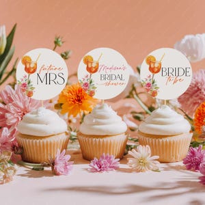 Könnte beinhalten: Drei Cupcakes mit weißem Zuckerguss und dekorativen Tortenauflegern. Die Aufleger zeigen eine Cocktail-Illustration, florale Akzente und den Text "future MRS", "Madison's Bridal Shower" und "BRIDE to be". Die Cupcakes sind von Blumen umgeben.