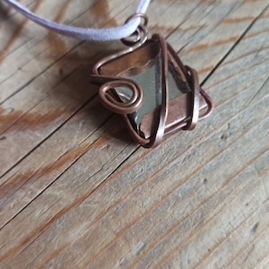 Pode incluir: Um pingente envolto em fio de cobre com uma pedra de vidro transparente. O pingente está preso a um cordão branco.