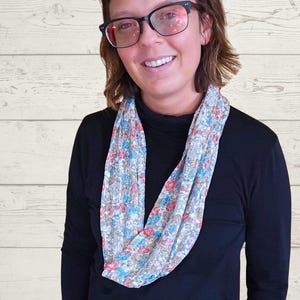 May include: A floral print infinity scarf in shades of blue, red, and beige. The scarf is draped around the neck, and the person is wearing a black long-sleeve shirt and glasses.