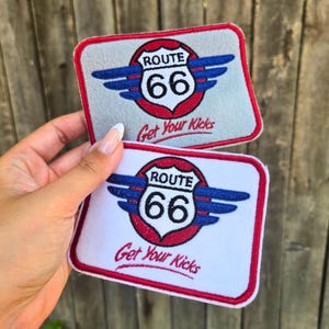 May include: Two rectangular embroidered patches featuring the Route 66 shield logo with blue wings and a red circular background. The text Route 66 and Get Your Kicks is stitched in black. One patch has a light gray base, and the other has a white base.