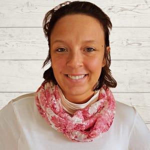 May include: A woman wearing a white turtleneck shirt and a pink and white floral print infinity scarf. The scarf is draped around the neck. The background is a white wood panel.