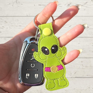 May include: A green alien keychain with a pink belt and black eyes is attached to a silver key fob. The alien keychain is made of stitched fabric and has a yellow button detail. The background is a light wood grain.