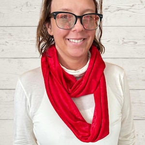 May include: A bright red infinity scarf made of a soft, velvety material. The scarf is draped around the neck and shoulders, creating a loop. The person in the image is wearing a white long-sleeved shirt and black glasses.